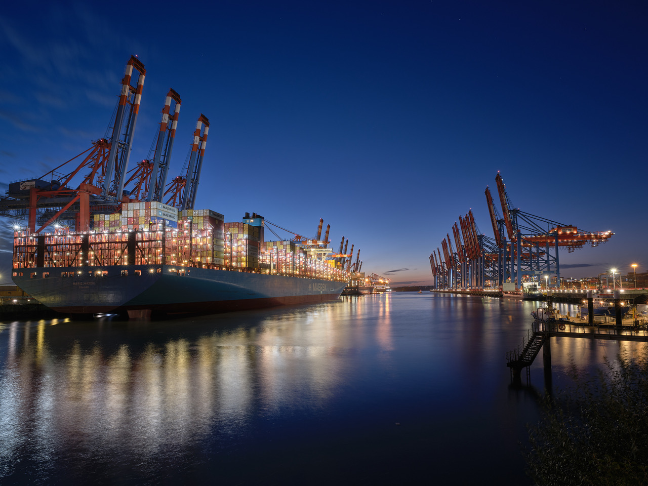 Frachtschiff bei Nacht, ruhig, blaues Wasser, leuchtende Krane und Container